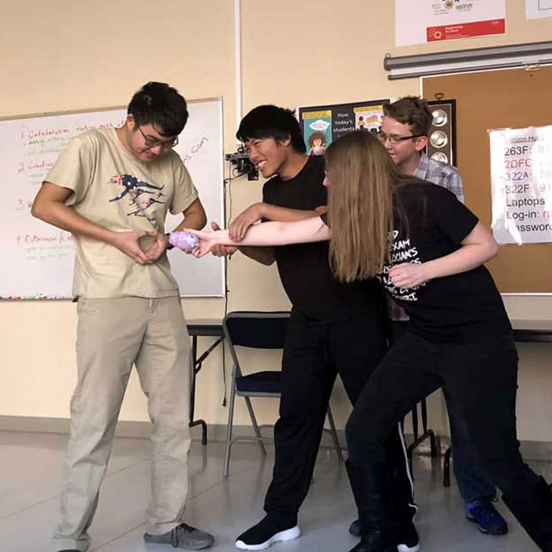 A group of four people are humorously interacting, with one person on the left holding a rubber chicken while the others reach towards it, standing in a classroom setting.