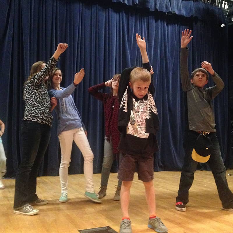 A group of children poses on a stage with blue curtains. They strike various playful and expressive poses, standing on a wooden floor.