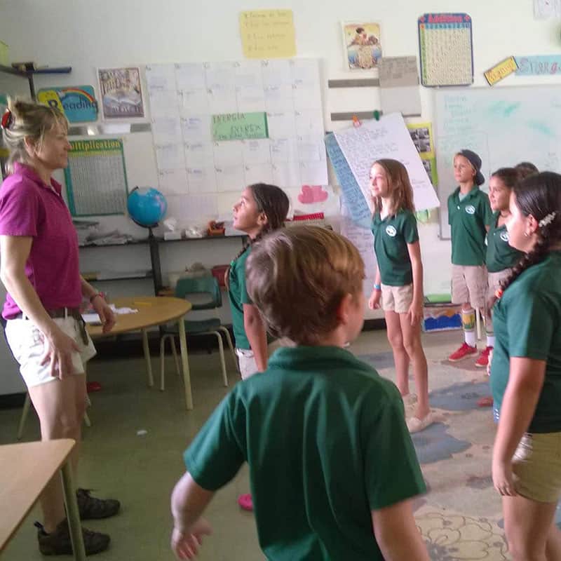 A teacher in a pink shirt leads a group of young students in a classroom. The students are standing and appear attentive, wearing matching green uniforms. The room is decorated with educational posters.